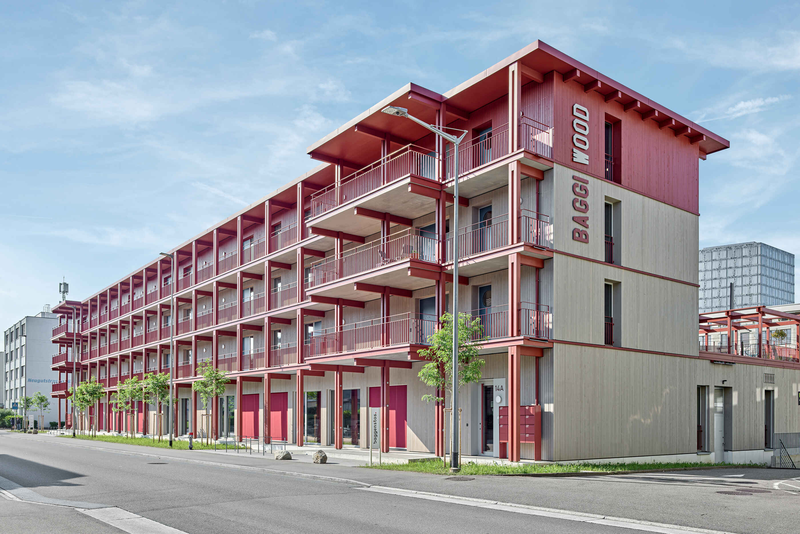 Aussenansicht des mehrstöckigen Baggiwood-Gebäudes mit rot-holzerner Fassade unter blauem Himmel