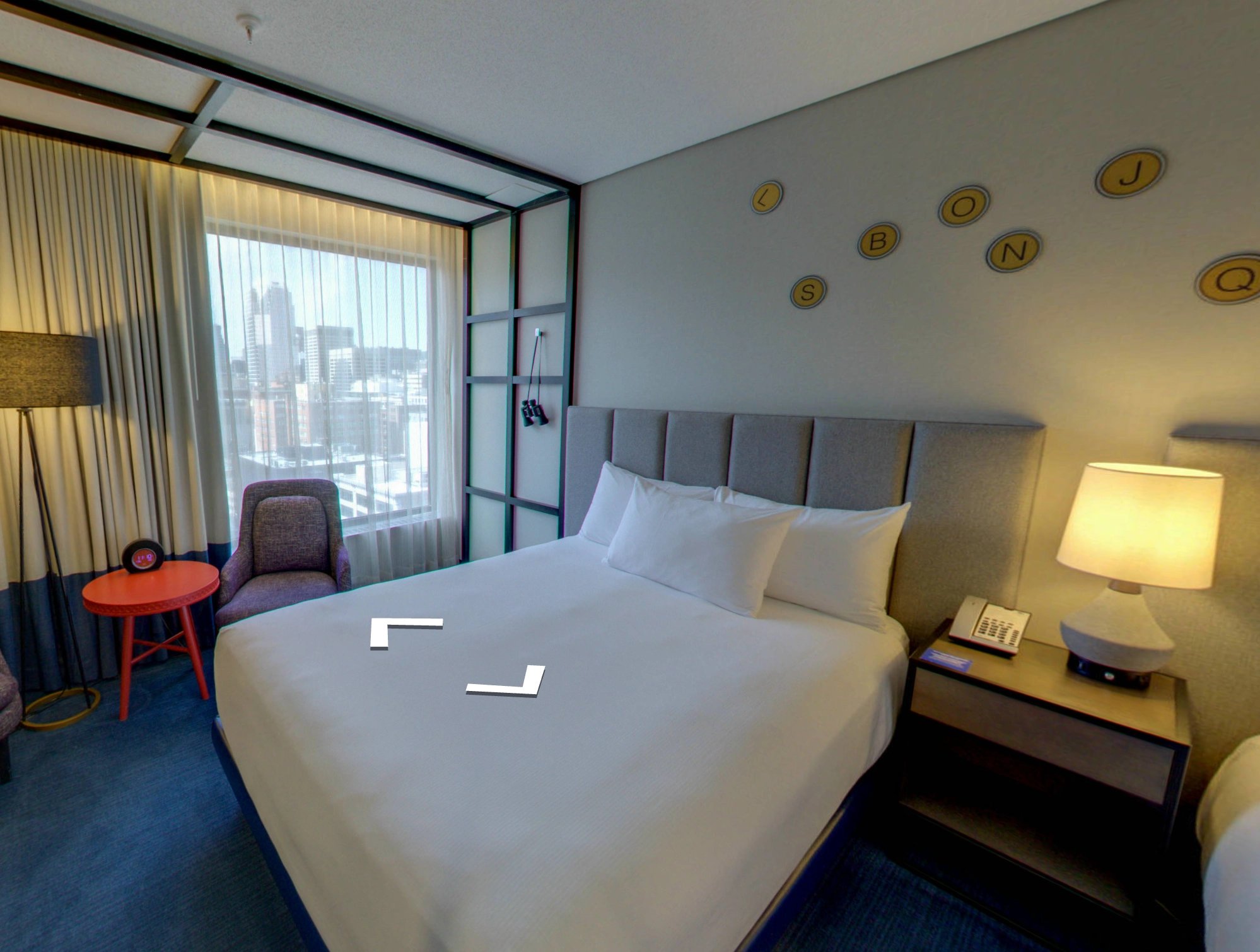 Modern hotel room with a large bed, gray upholstered headboard, bedside table with lamp and phone, purple chair, red round table, and a city view through a large window with sheer curtains.