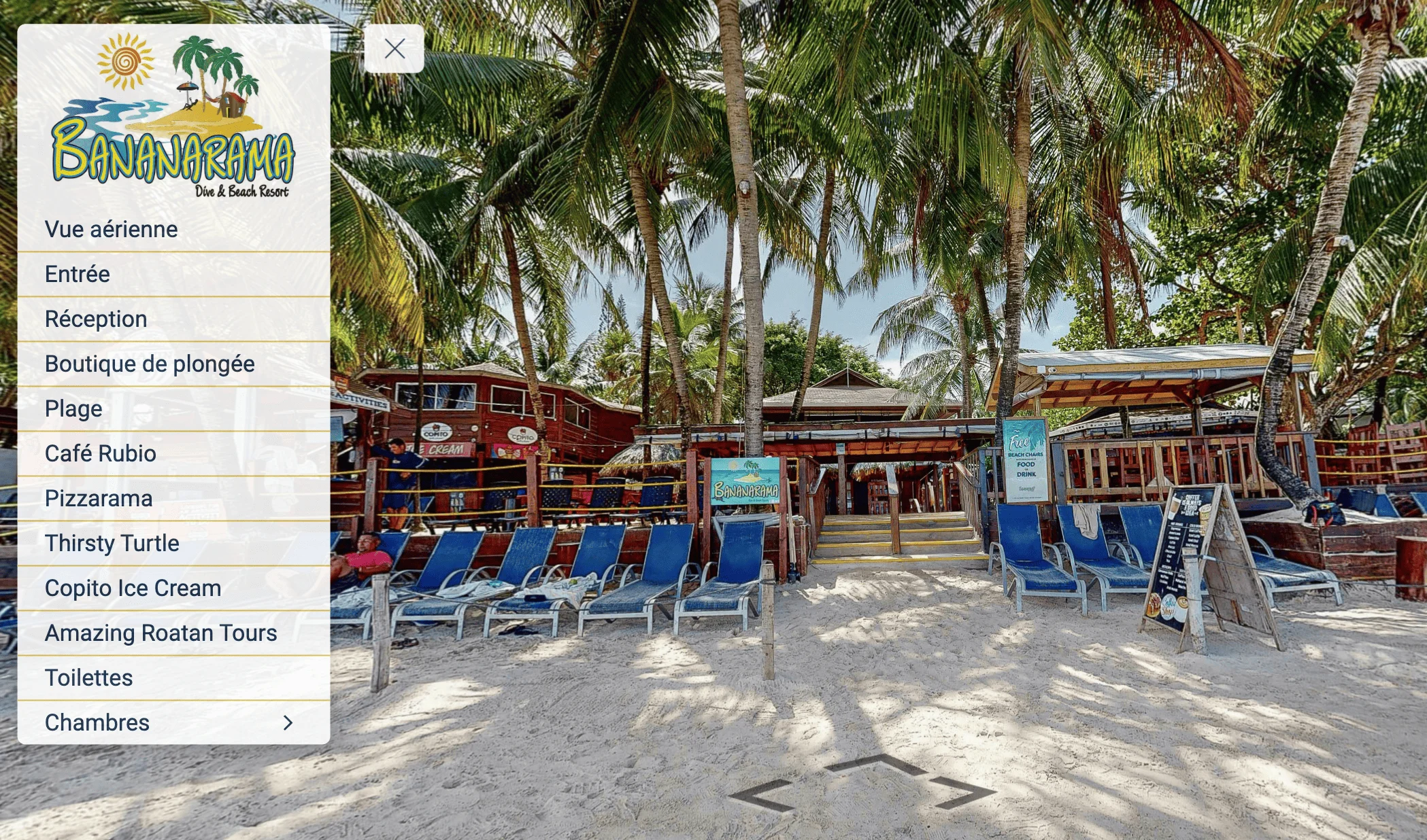 Sandy beach with blue lounge chairs under palm trees and a wooden resort building with signage for Bananarama Dive & Beach Resort.