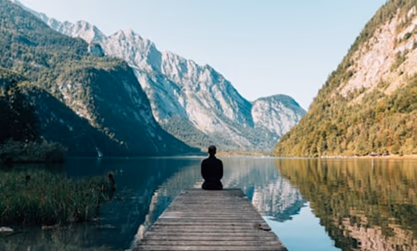 Person sitting at the end of a wooden dock overlooking a calm lake surrounded by mountains.