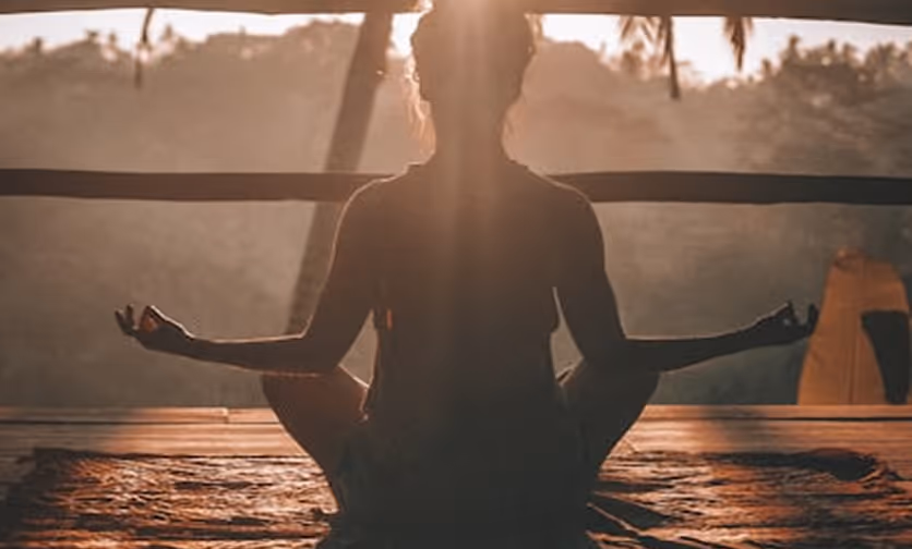 Silhouette of a person meditating cross-legged indoors with sunlight streaming through a window.