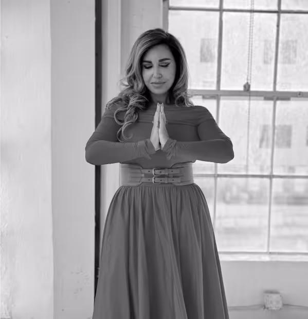 A woman in a floral dress sitting cross-legged on a patterned blanket by a river, meditating with her eyes closed and hands resting on her knees, with a cup in front of her.