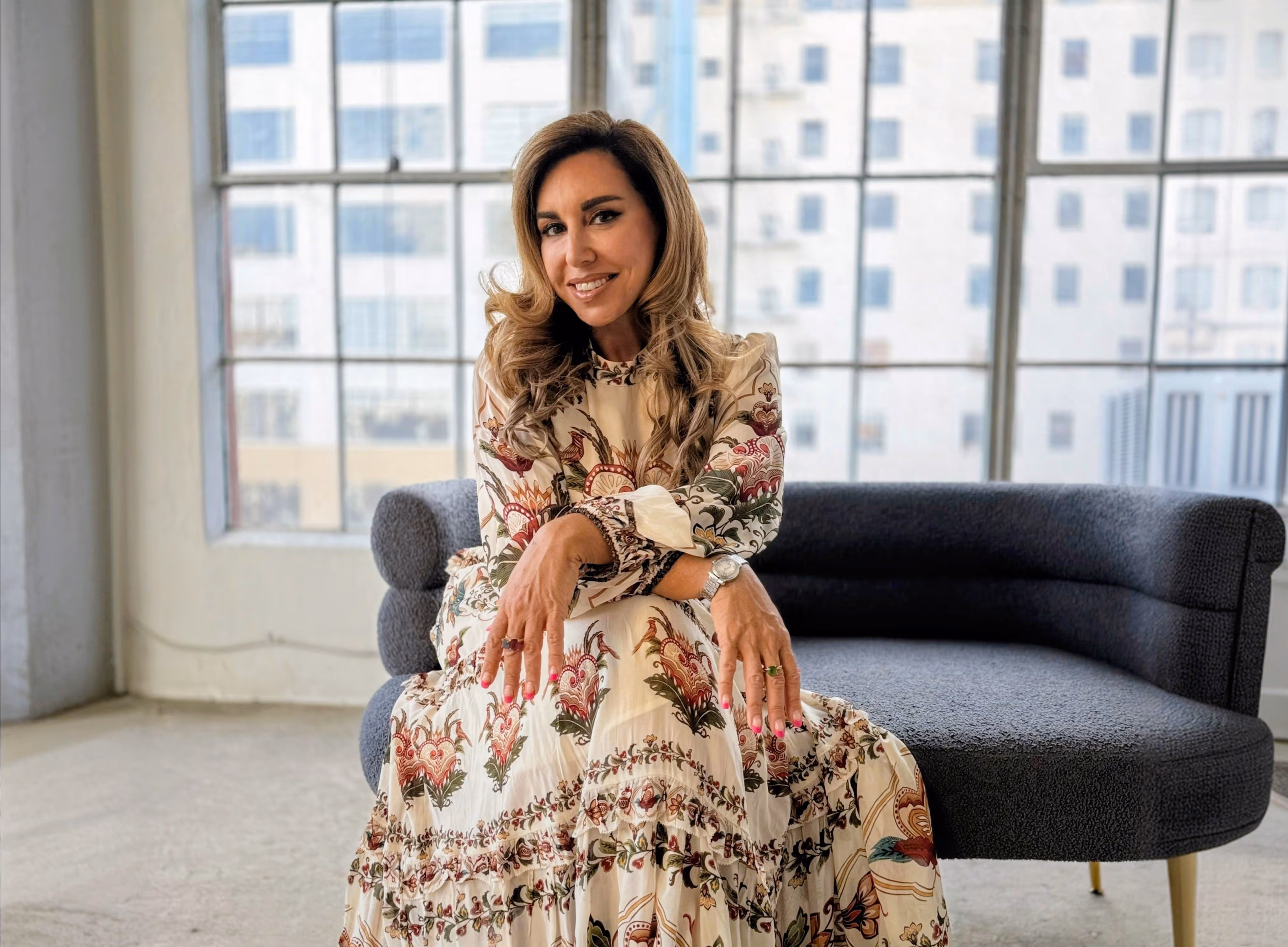 Woman with long wavy hair wearing a floral patterned dress sitting on a modern gray couch in front of large windows.