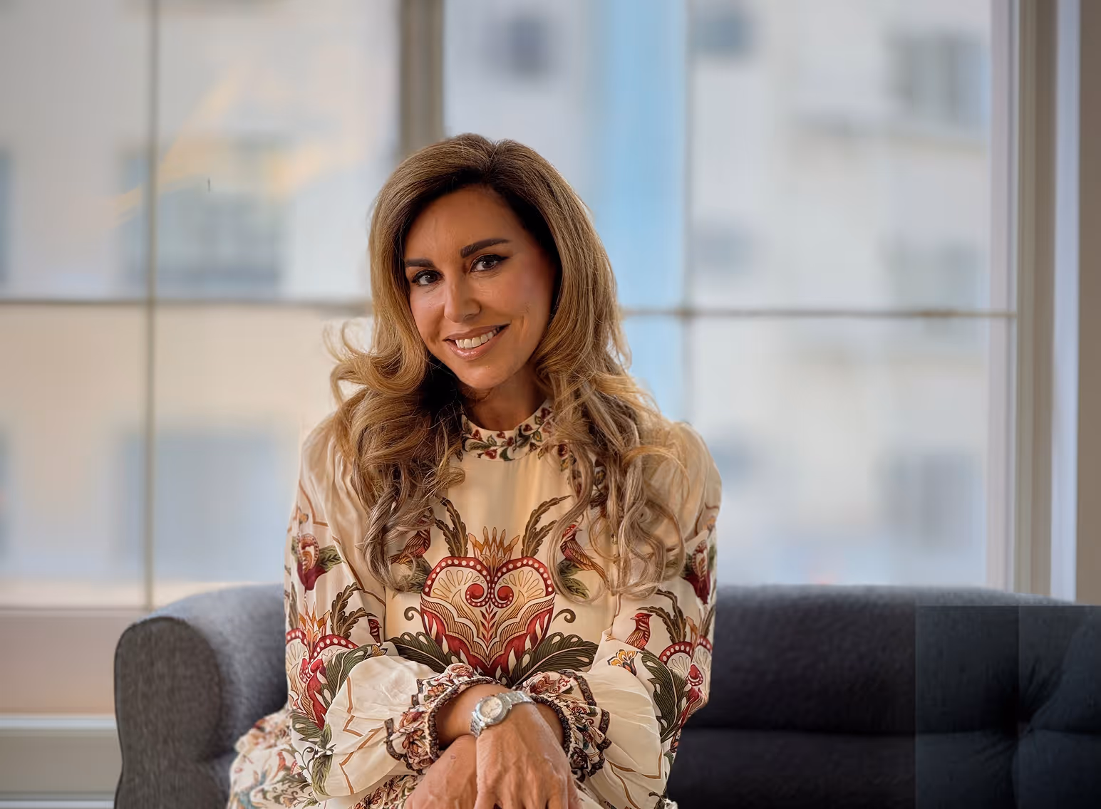 Smiling woman with long wavy hair wearing a patterned blouse sitting on a gray couch in front of a large window.