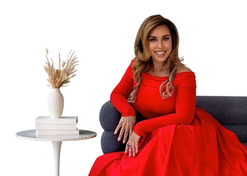 Smiling woman in a long red dress sitting on a gray sofa next to a table with stacked white books and a vase with dried plants.