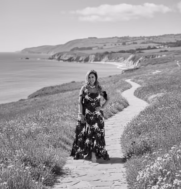 Woman in a floral dress standing on a winding stone path surrounded by grassy fields with a coastal cliff and ocean in the background.