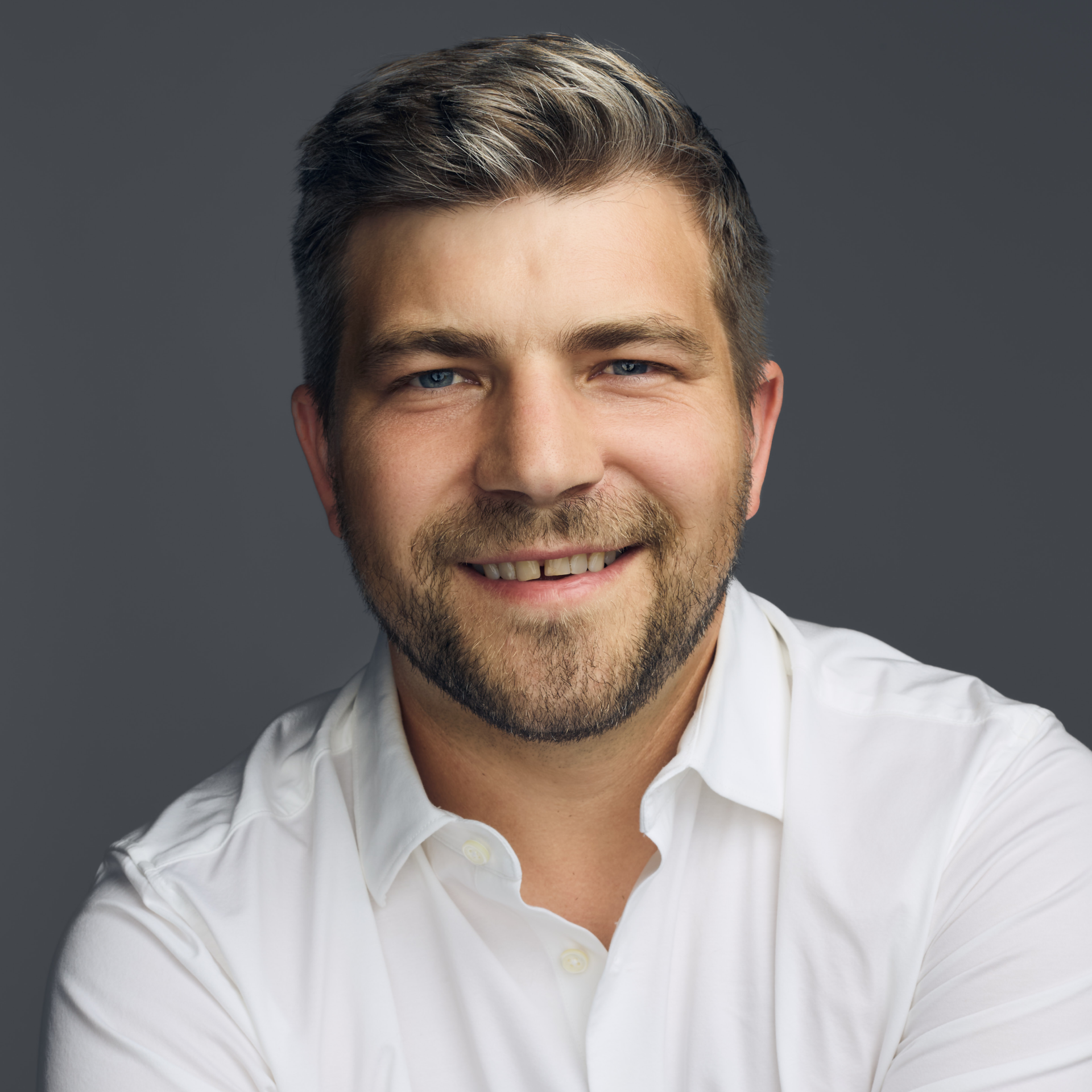 Smiling man with blue eyes, beard, and short hair wearing a white shirt against a gray background.