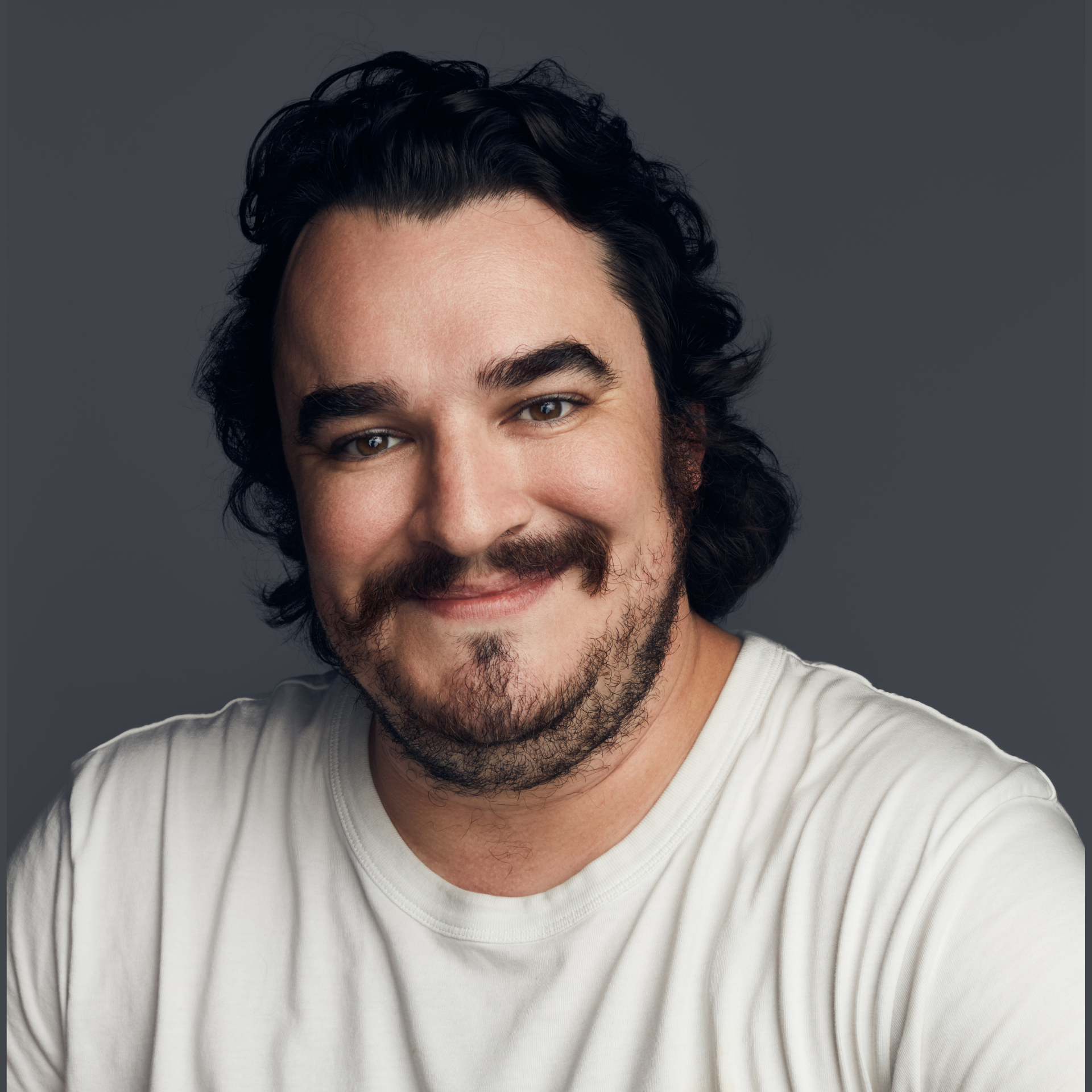Smiling man with dark curly hair and beard wearing a white T-shirt against a gray background.