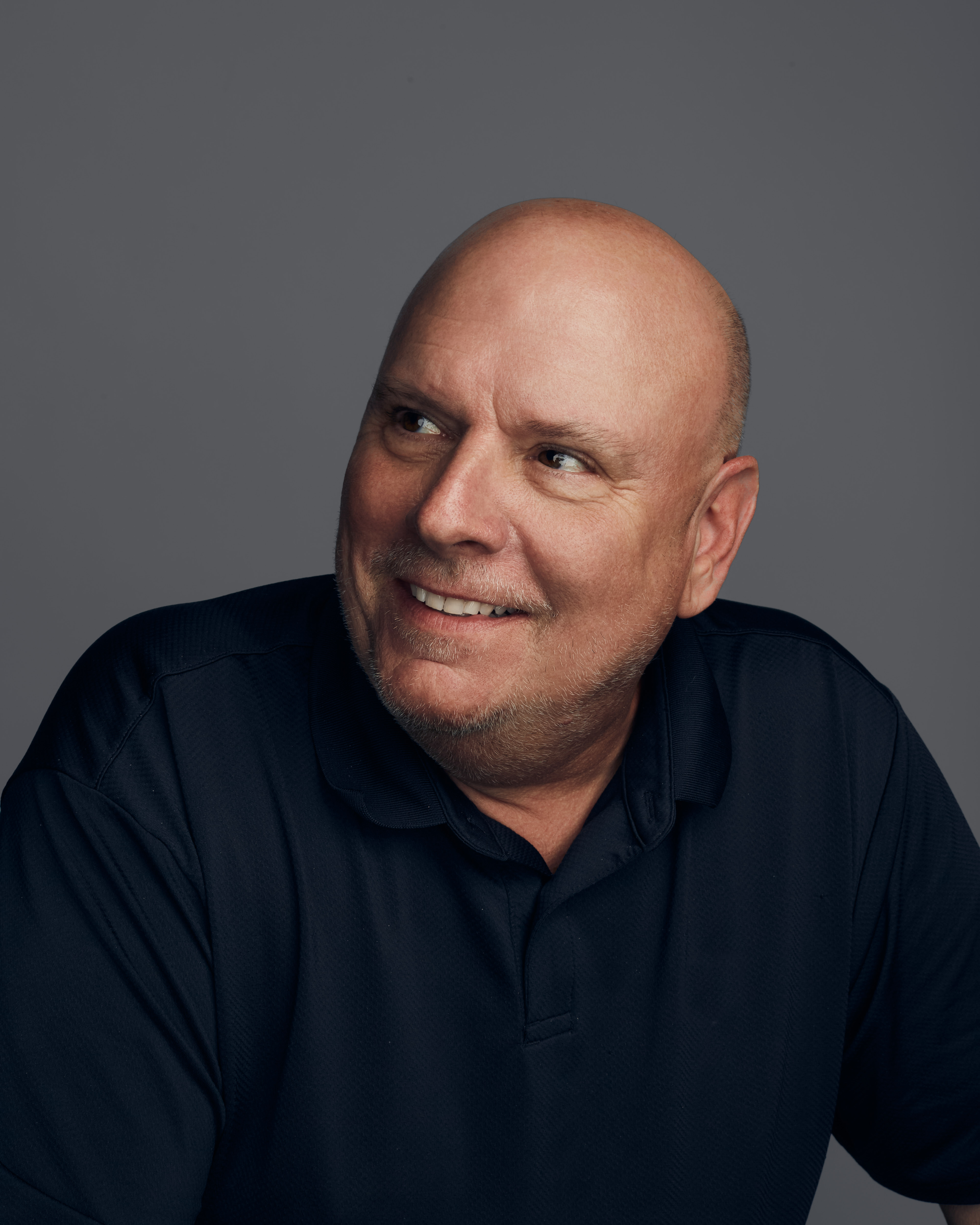 Smiling bald man wearing a dark polo shirt looking to his left against a gray background.