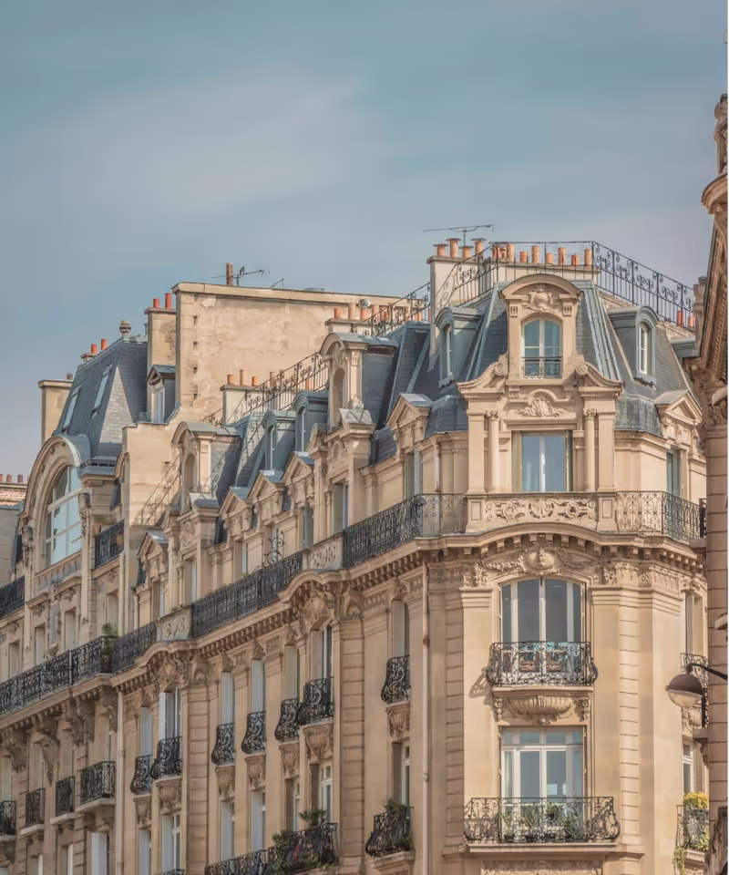 Façade d'un immeuble haussmannien parisien avec balcons en fer forgé et toits mansardés sous un ciel bleu clair.