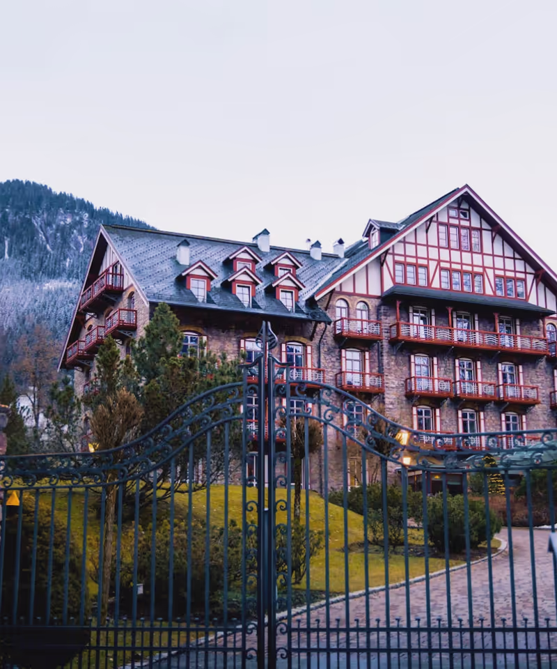 Grand bâtiment en pierre avec balcons rouges, entouré d'un jardin et d'une grille en fer forgé, avec montagne en arrière-plan.
