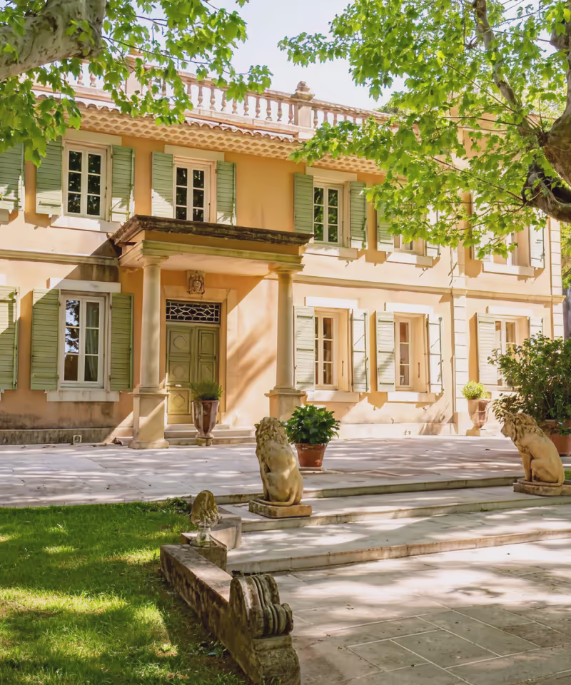 Façade d'une maison beige avec volets verts, entrée avec colonnes et statues de lions en pierre sur une terrasse ensoleillée.