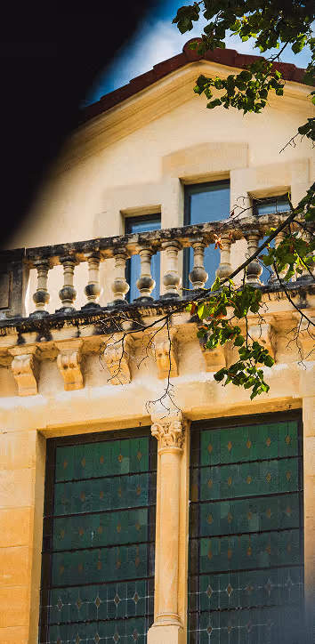 Façade d'un bâtiment ancien en pierre avec balustrade, colonnes et fenêtres à vitraux verts, partiellement encadrée par des branches d'arbre.