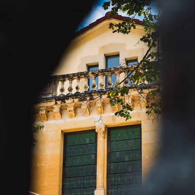 Façade d'un bâtiment ancien en pierre avec balustrade, colonnes et fenêtres à vitraux verts, partiellement encadrée par des branches d'arbre.