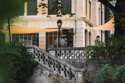 Terrasse extérieure avec balustrade en pierre, lampadaire et grandes baies vitrées sur un bâtiment en pierre claire, entouré de végétation.