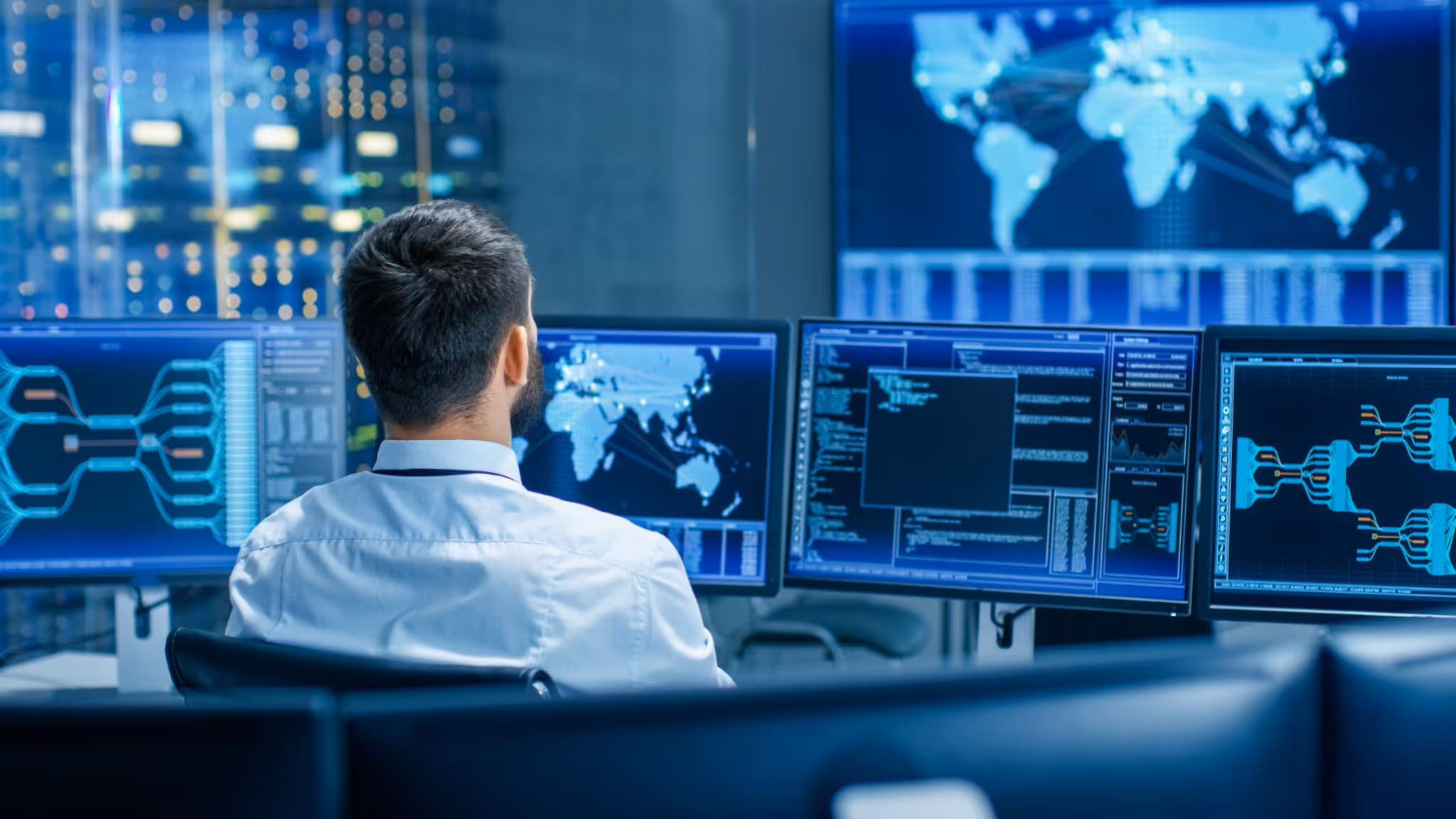 Man in white shirt monitors multiple computer screens with global maps and data analytics in a control room.