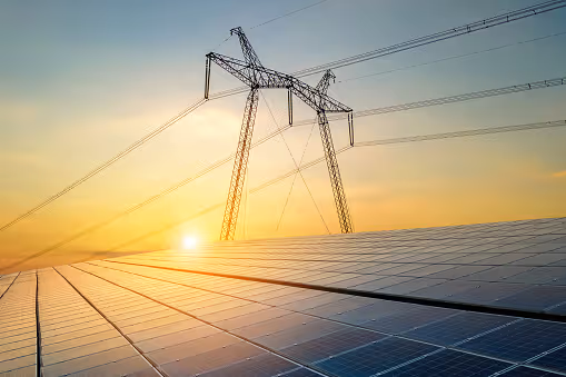 Solar panels in foreground with electricity transmission tower and power lines at sunset.