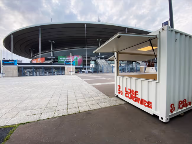 Kiosque blanc avec inscription rouge ouvert devant un grand stade arrondi sous un ciel nuageux.