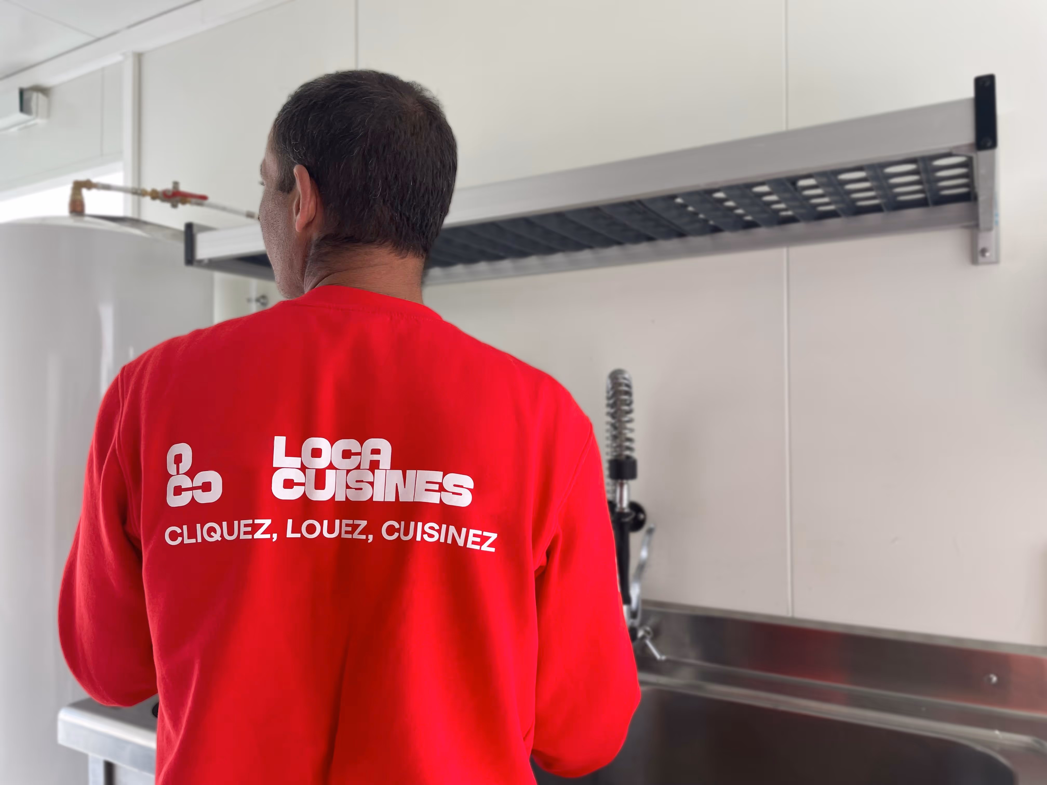Homme de dos portant un t-shirt rouge avec le texte 'Loca Cuisines, Cliquez, Louez, Cuisinez' dans une cuisine professionnelle avec un évier en acier inoxydable.