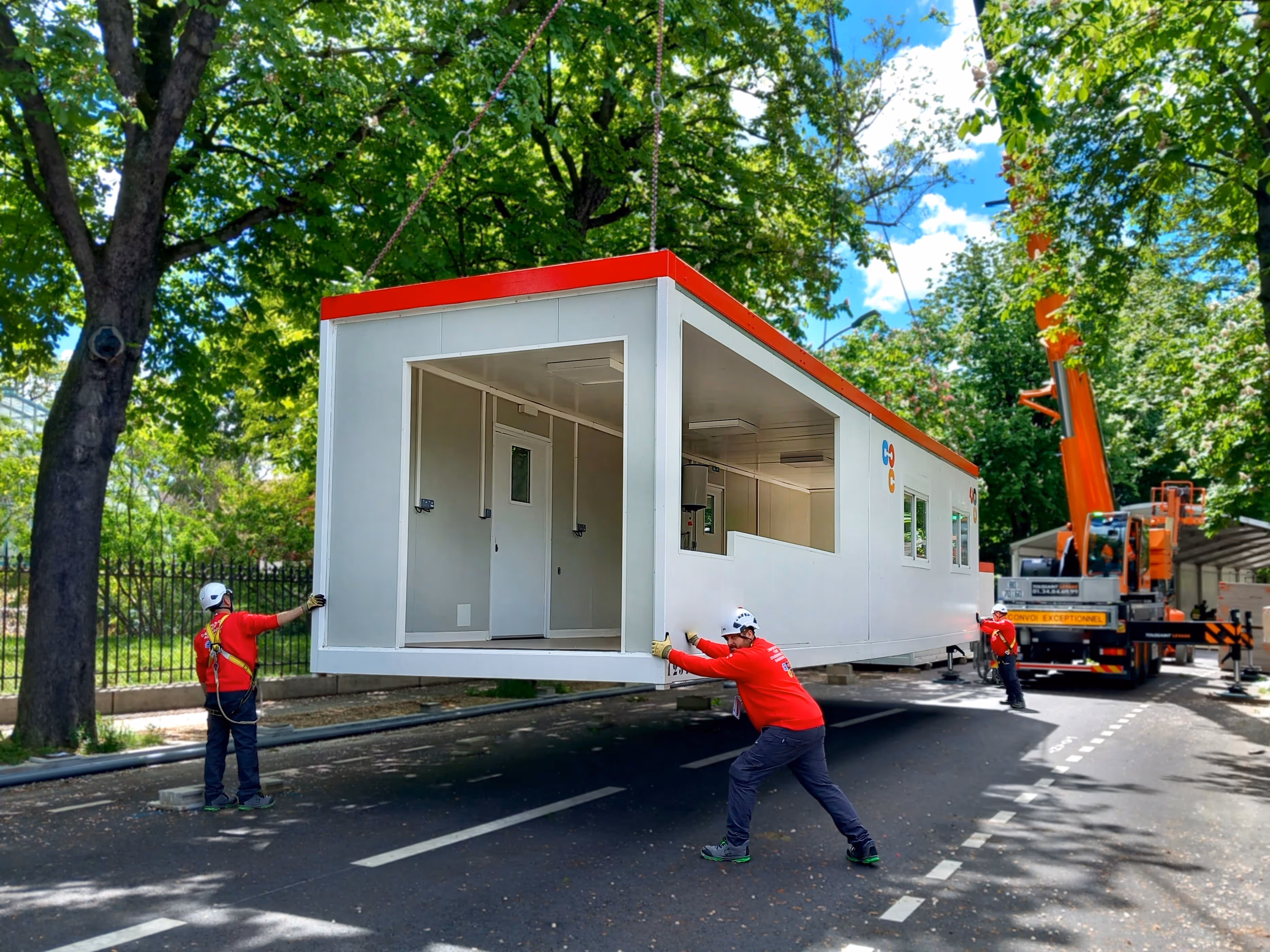 Trois ouvriers en casque et tenue rouge déplacent un module préfabriqué suspendu par une grue orange sur une route bordée d'arbres verts.