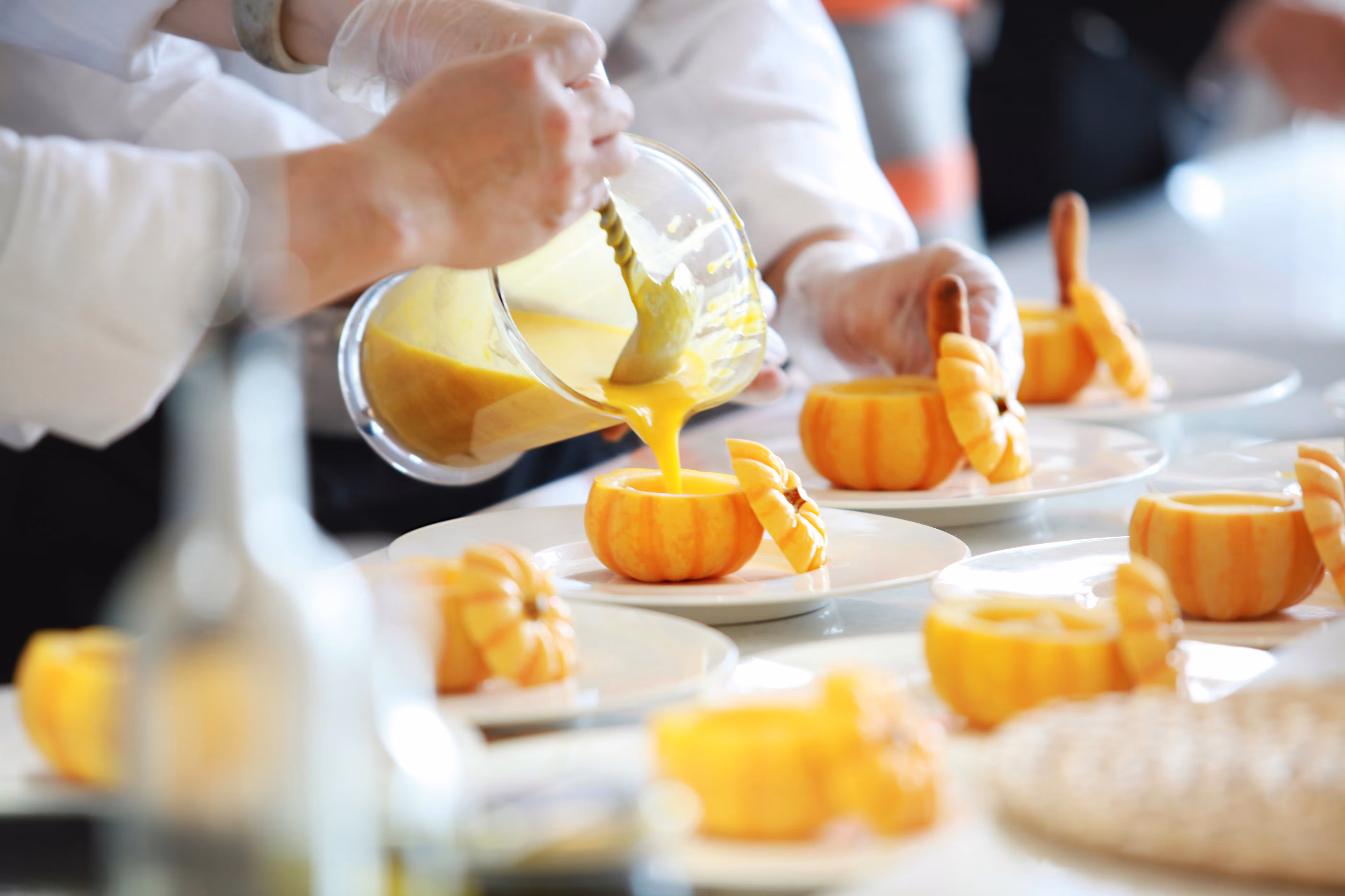 Versement de soupe jaune dans de petites citrouilles évidées disposées sur des assiettes blanches.