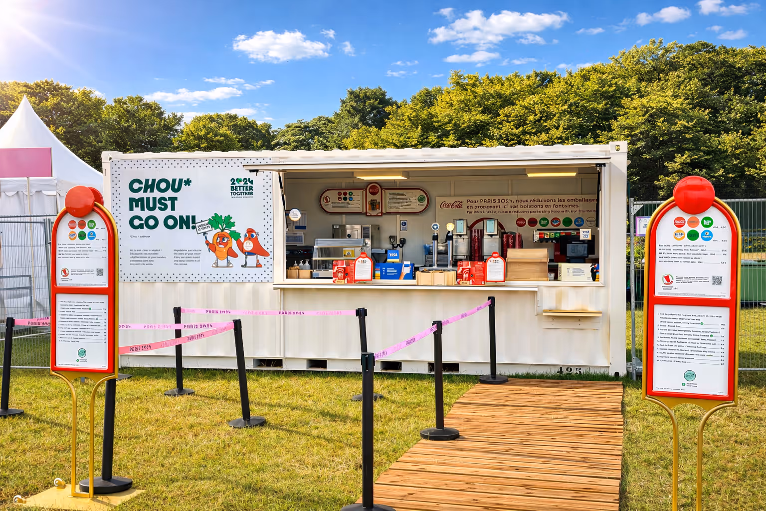Kiosque alimentaire en plein air avec deux panneaux de menu rouge et jaune sur l'herbe, ciel bleu et arbres en arrière-plan.