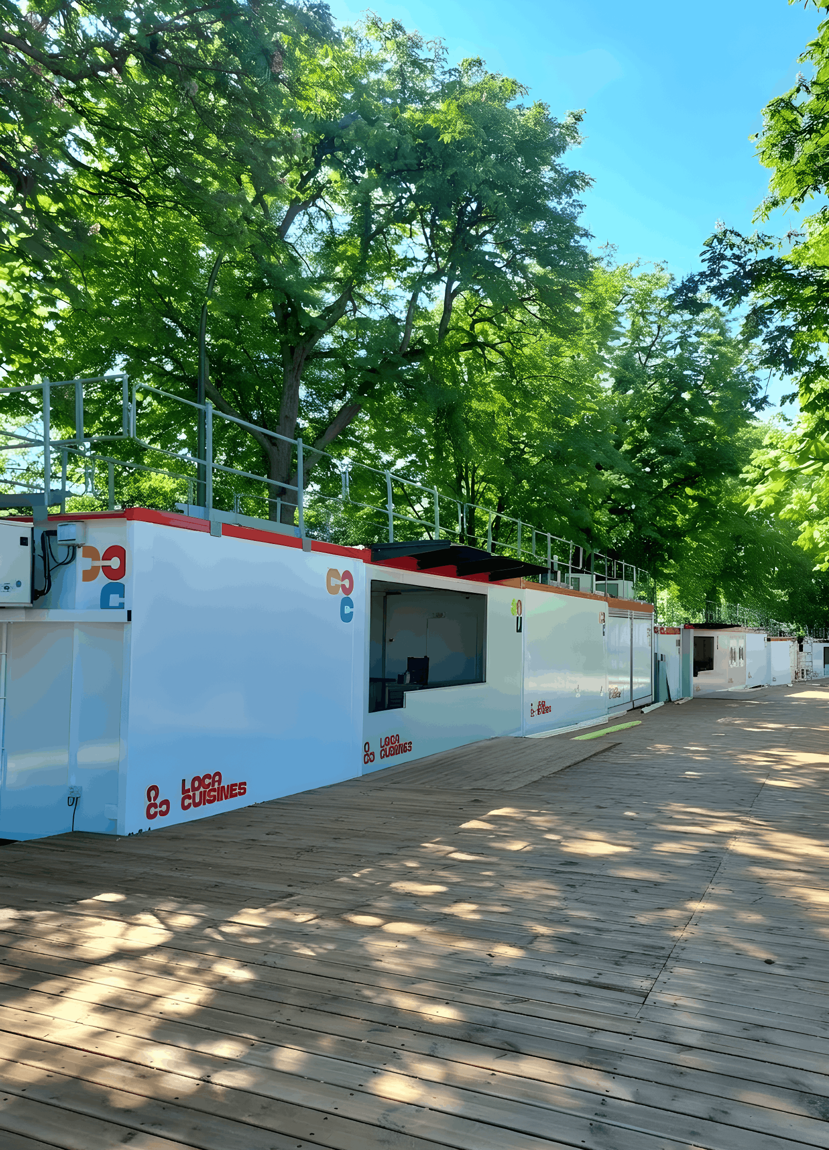 Conteneurs blancs Locacuisines  alignées sur une passerelle en bois sous de grands arbres verts.