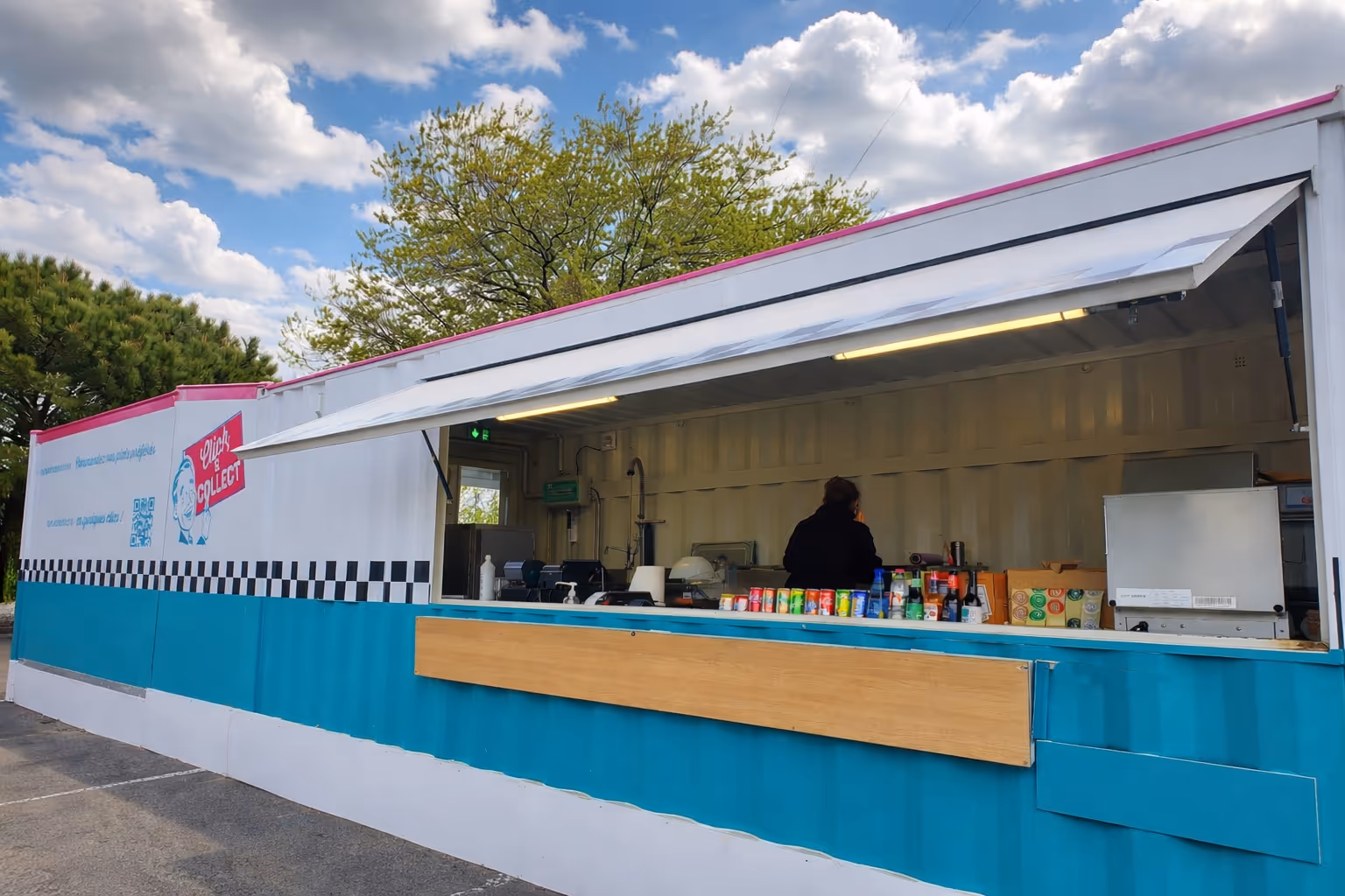Un stand de restauration mobile bleu et blanc avec un auvent ouvert, une personne à l'intérieur et une rangée de boissons en canettes sur le comptoir.