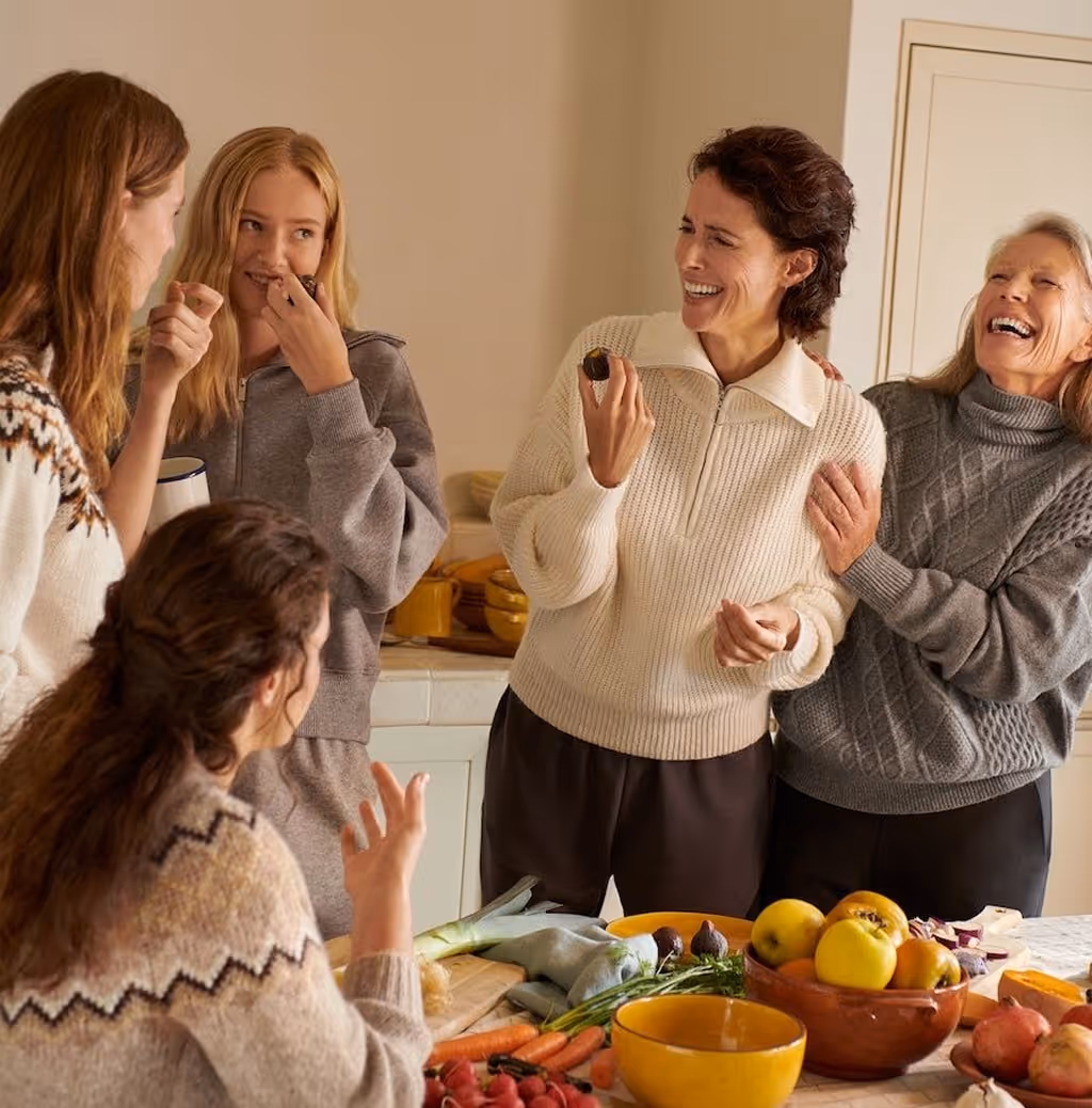 Vijf lachende vrouwen in warme truien praten en delen eten rond een tafel met fruit en groenten in een huiselijke keuken.