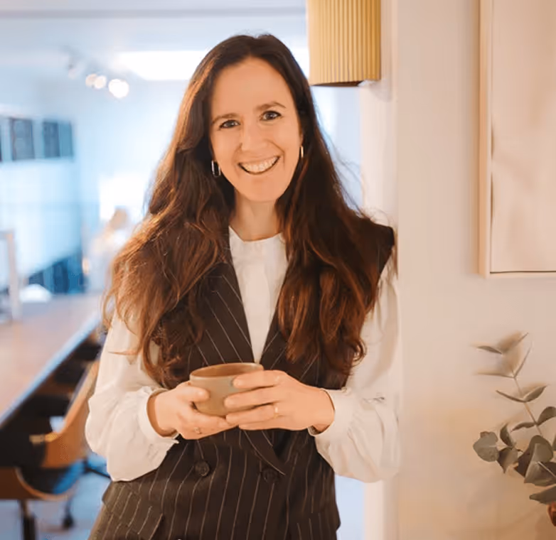 Vrolijke vrouw met lang bruin haar en een gestreept zwart vest, die een kopje vasthoudt in een lichte, moderne kamer.