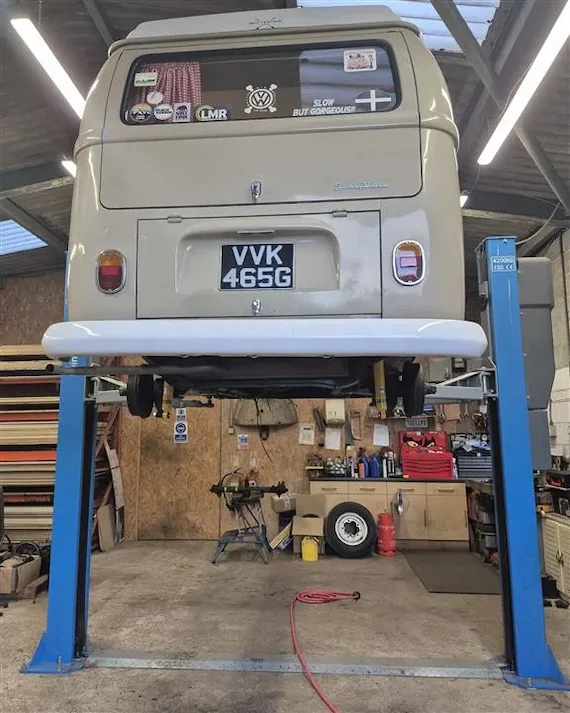 Rear view of a beige classic Volkswagen camper van with license plate VVK 465G lifted on a blue vehicle hoist inside a garage.