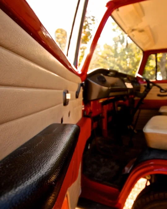 Interior view of a vintage red vehicle showing the door panel, black armrest, dashboard, and beige seats with sunlight coming through the windows.