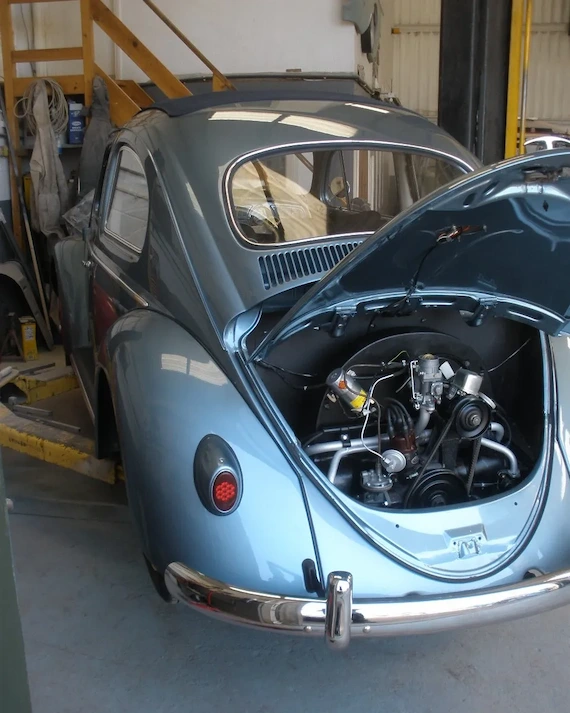 Rear view of a light blue vintage Volkswagen Beetle car with its engine compartment open inside a garage.