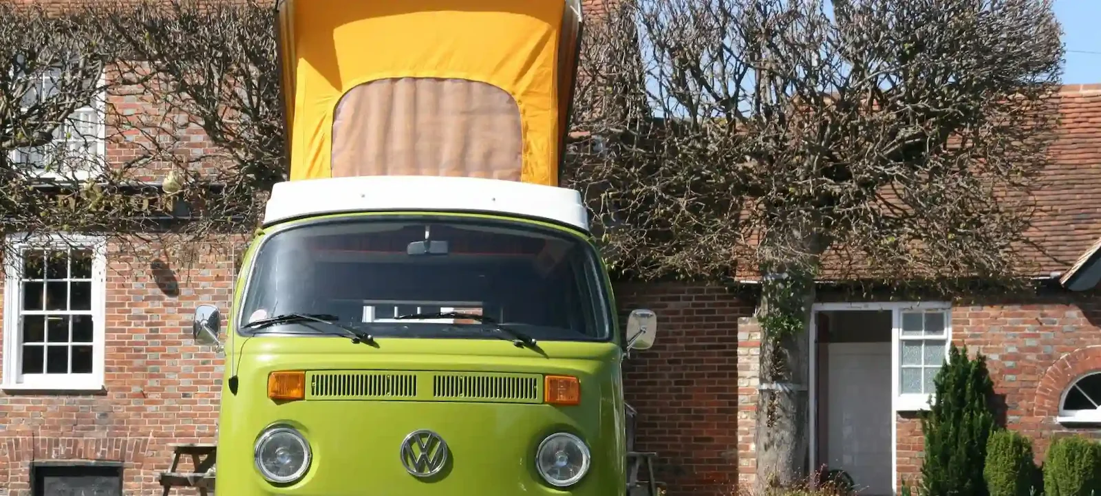 Front view of a green vintage Volkswagen camper van with a yellow pop-up roof parked in front of a brick house with leafless trees.