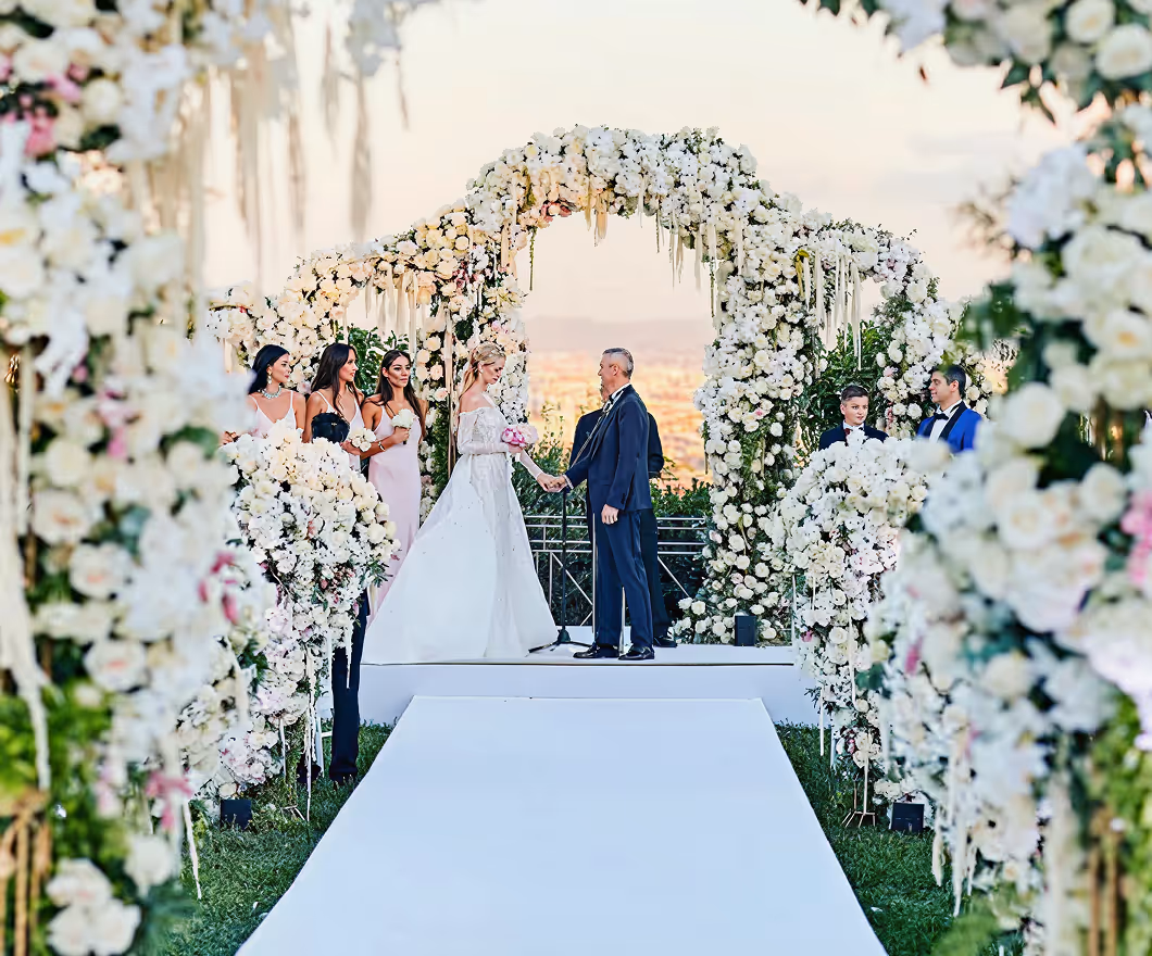 Bride and groom holding hands at an outdoor wedding altar adorned with abundant white flowers and surrounded by bridesmaids and groomsmen.