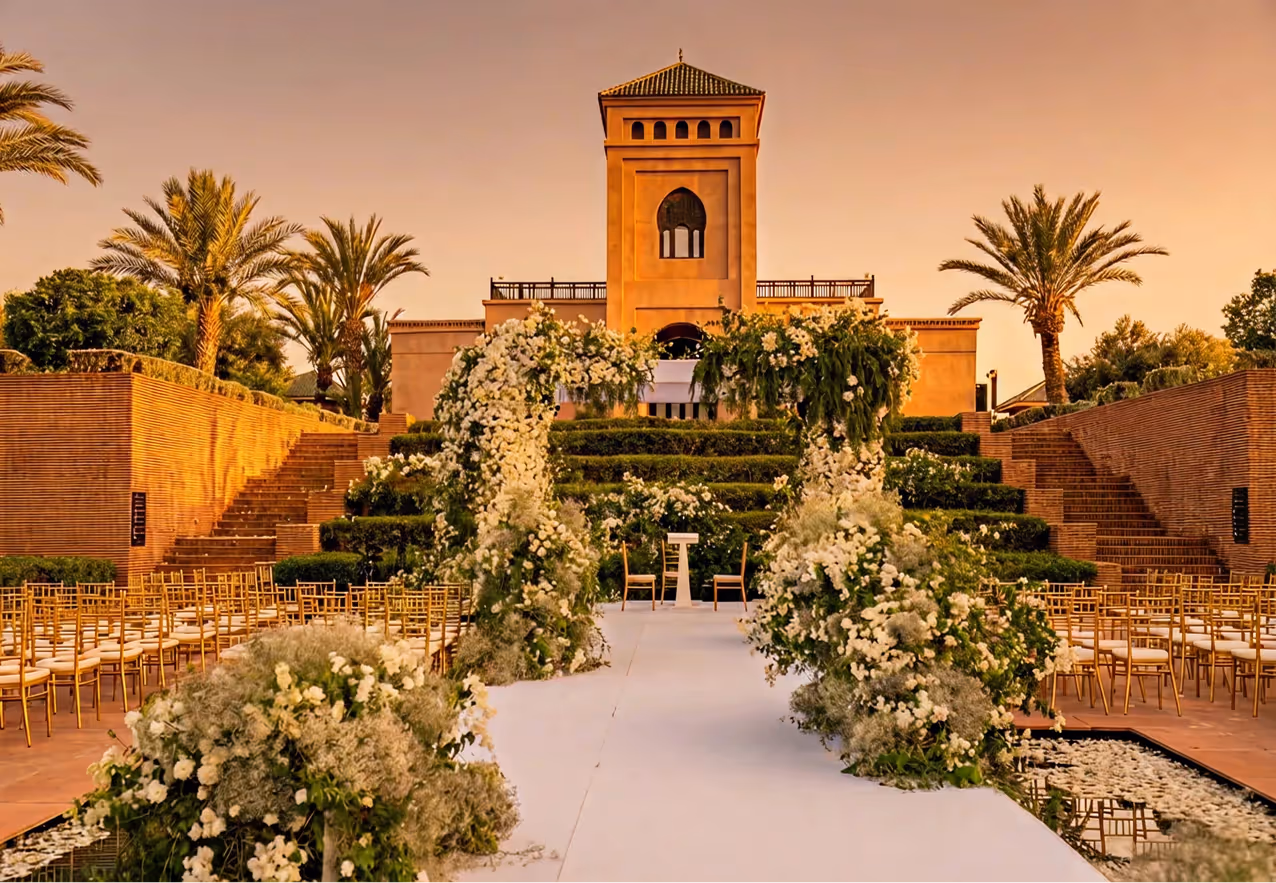 Elegant outdoor gathering with people seated at white tables in front of a historic white building with ornate architecture and potted plants.