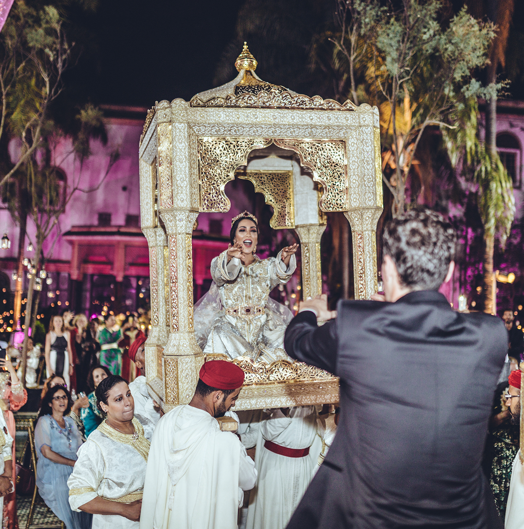 A bride dressed in ornate white and gold sits joyfully in a decorated golden palanquin carried by men in traditional attire during a nighttime wedding celebration.