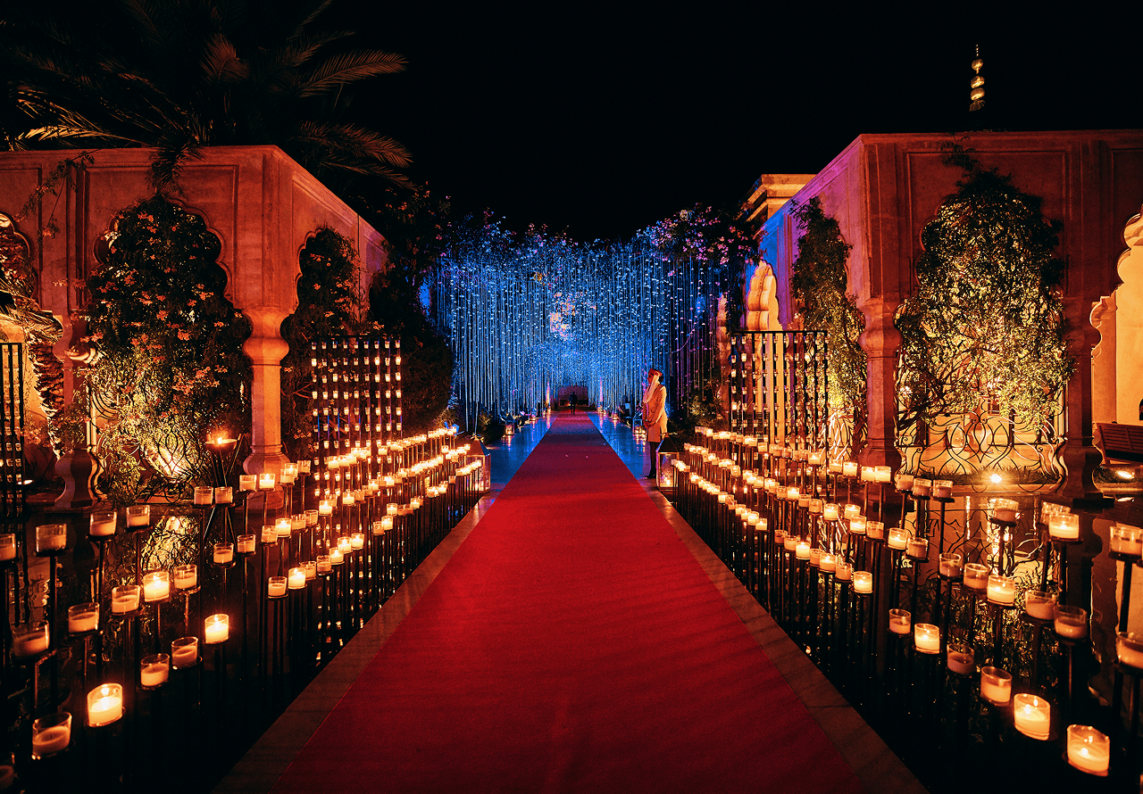 Pathway with lit candles on both sides, red carpet, and blue hanging lights at night in a decorative garden setting.