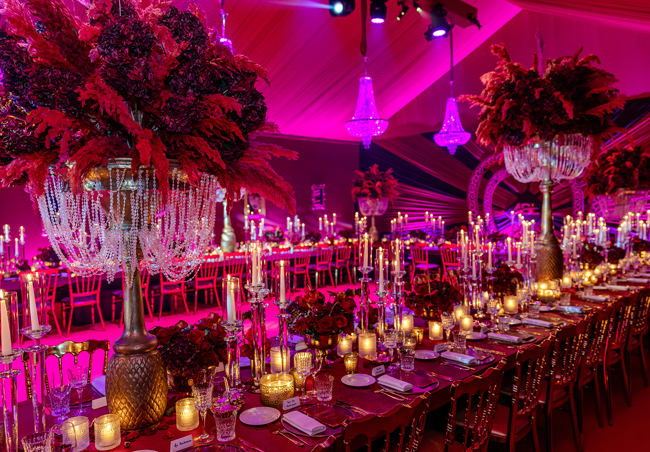 Elegantly decorated banquet hall with long tables featuring red floral centerpieces, numerous lit candles, crystal glassware, and chandeliers under pink-purple lighting.