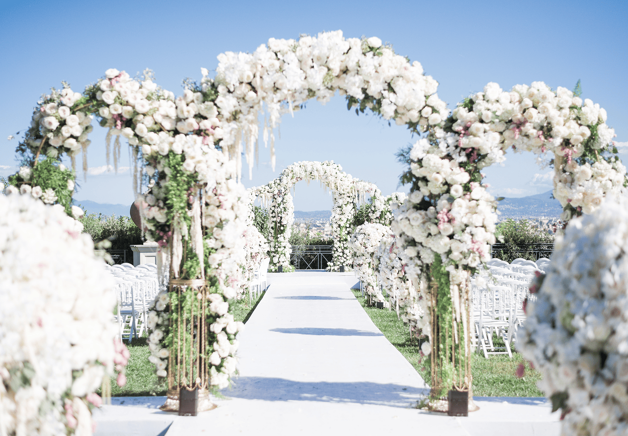 Elegant outdoor gathering with people seated at white tables in front of a historic white building with ornate architecture and potted plants.