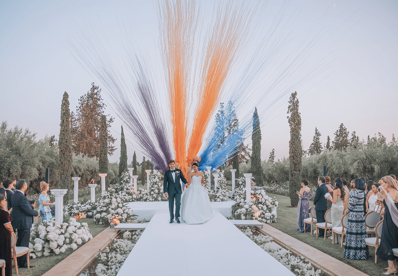Newlywed couple walking hand in hand down a white aisle during an outdoor wedding ceremony with colorful orange, purple, and blue streamers exploding behind them.