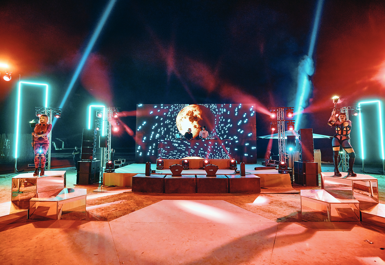 Outdoor concert stage at night with two DJs performing and two performers holding torches on mirrored platforms, illuminated by colorful lights.
