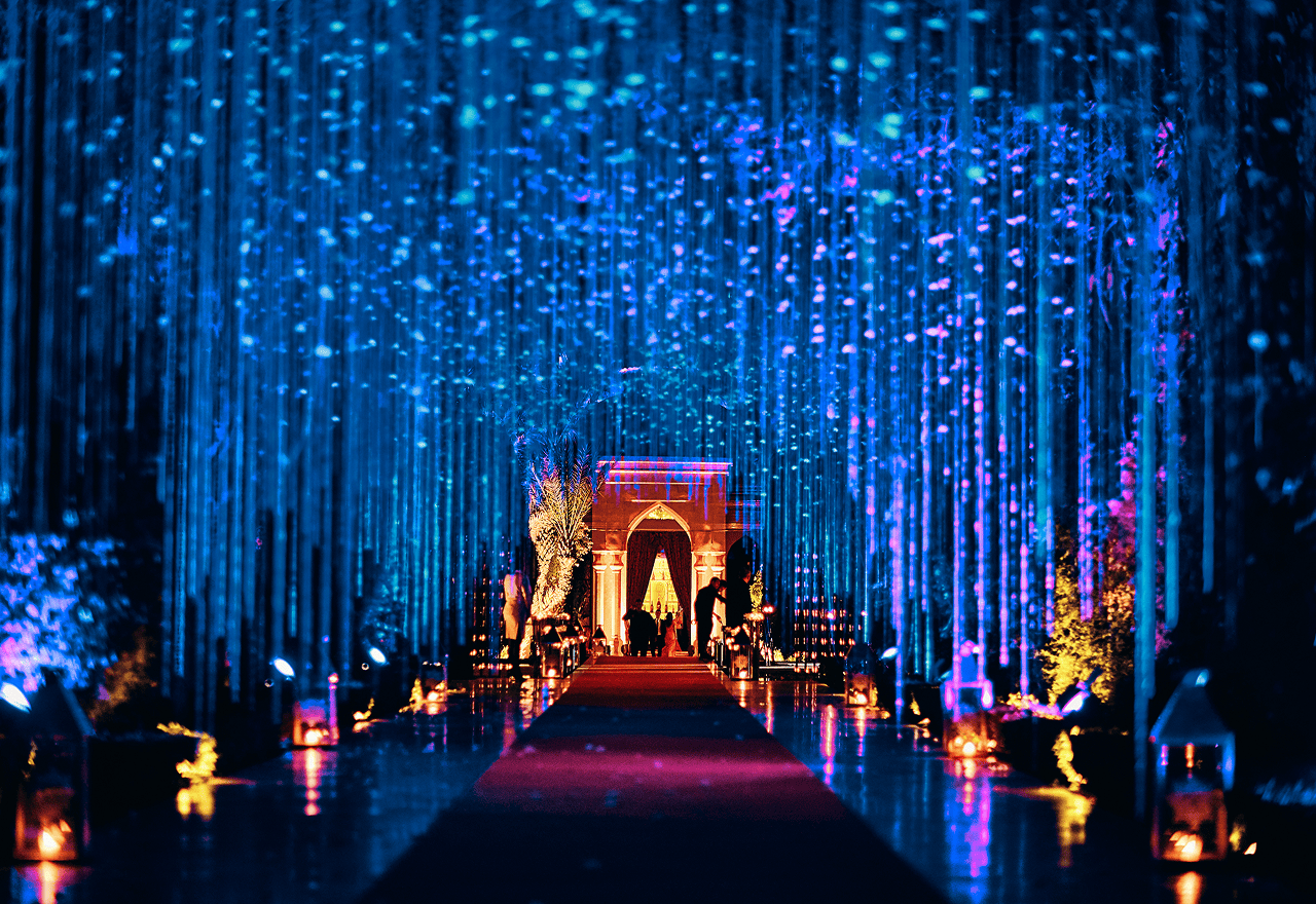 Illuminated walkway decorated with hanging blue lights and lanterns leading to an ornate archway with silhouettes of people.