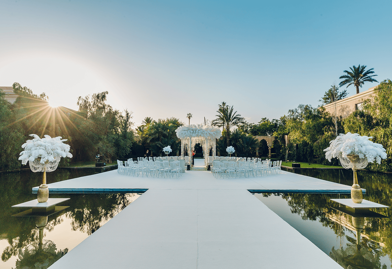 Outdoor wedding setup on a white platform over water with chairs arranged around a flower-adorned gazebo under a clear sky at sunset.