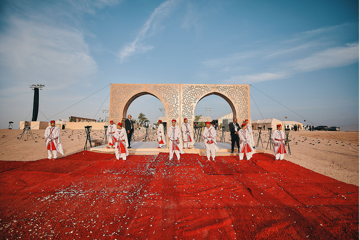 Outdoor wedding aisle decorated with white and pink floral arrangements and a flower-covered arch under a clear blue sky.