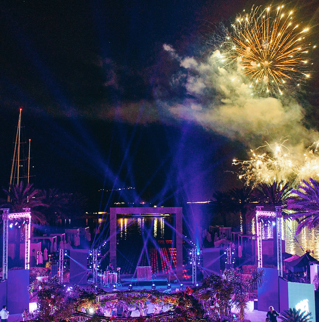 Nighttime outdoor event with stage lighting, palm trees, water reflections, and fireworks in the sky.