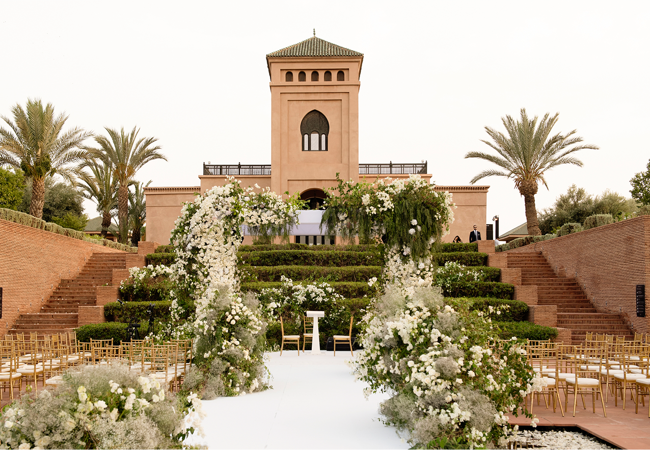 Elegant outdoor event with guests dining beside a large historic white building with ornate windows and greenery.
