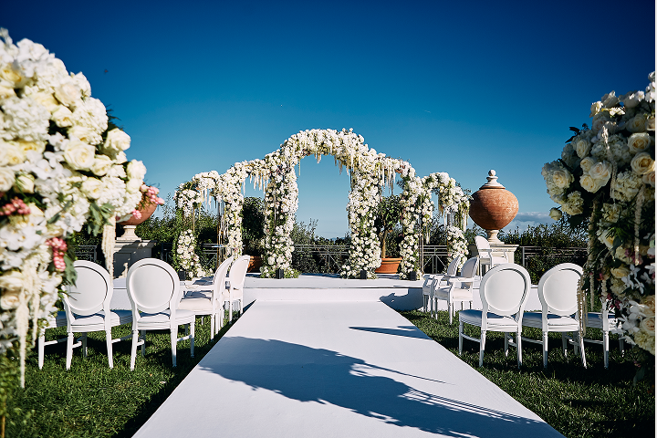 Outdoor wedding aisle decorated with white and pink floral arrangements and a flower-covered arch under a clear blue sky.