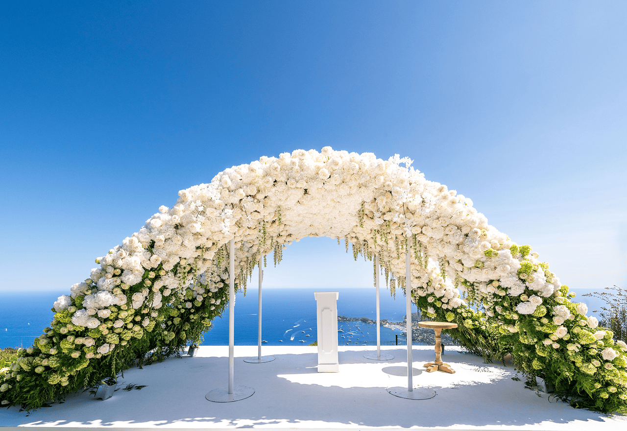 Elegant outdoor gathering with people seated at white tables in front of a historic white building with ornate architecture and potted plants.