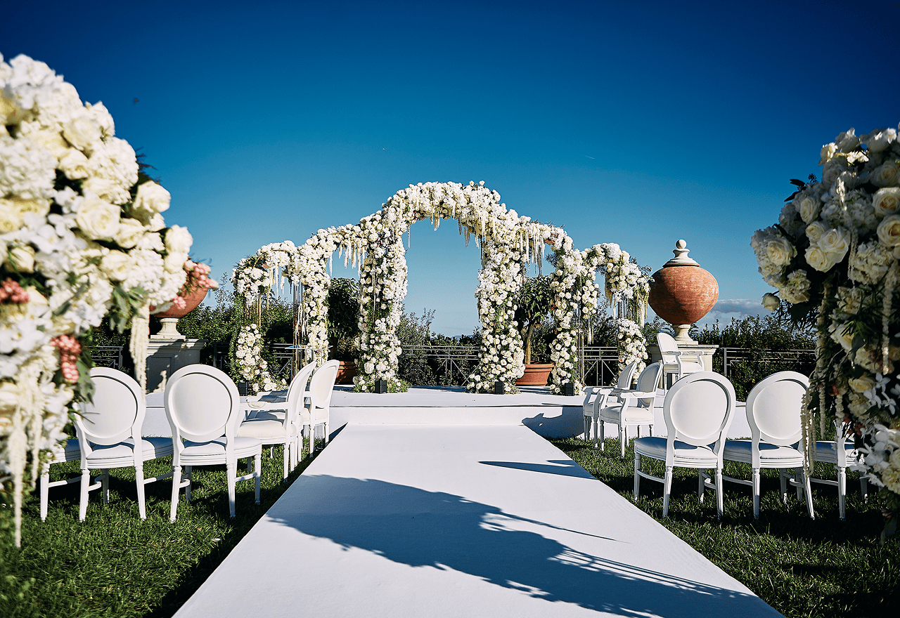 Elegant outdoor event with guests dining beside a large historic white building with ornate windows and greenery.